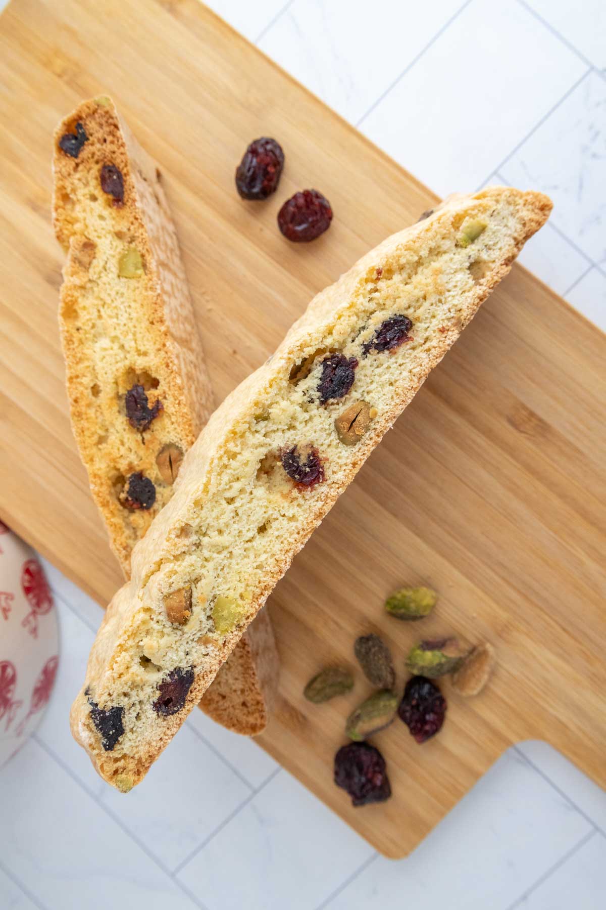 Two slices of biscotti with cranberries and pistachios on a wooden board, accompanied by scattered dried cranberries and pistachios.