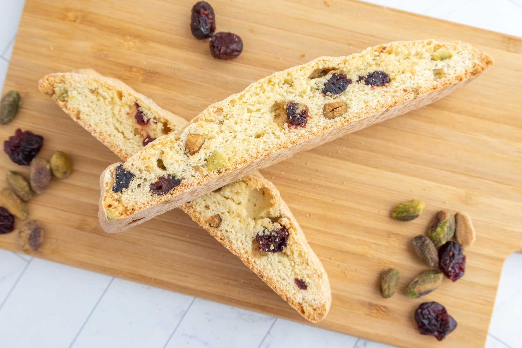 Two biscotti with pistachio and cranberry pieces on a wooden board, surrounded by loose pistachios and cranberries.