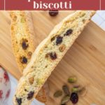 Two slices of cranberry pistachio biscotti on a wooden cutting board, surrounded by scattered pistachios and cranberries.