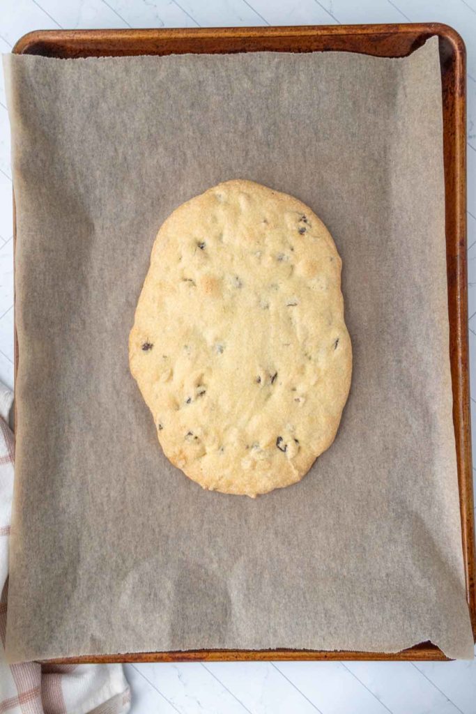 A large, flat cookie with chocolate chips on a parchment-lined baking sheet.