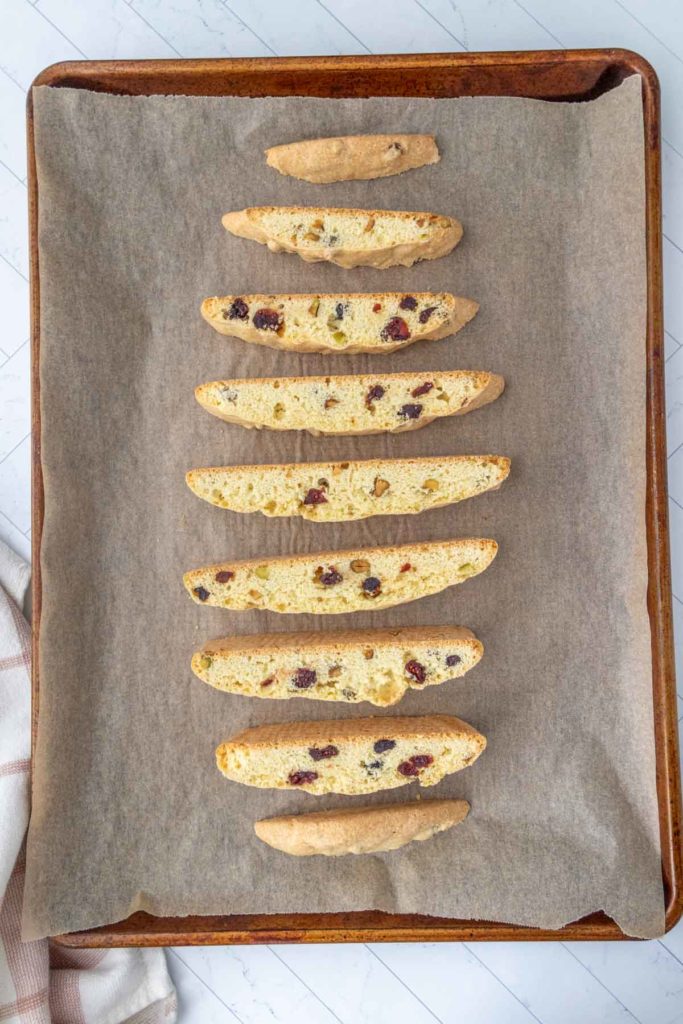 A baking sheet with nine slices of biscotti containing visible nuts and dried fruits, arranged in a line on parchment paper.