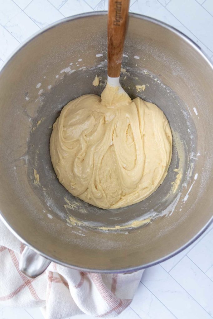 A metal mixing bowl containing a smooth, creamy batter with a wooden spatula. A kitchen towel is placed beside it.