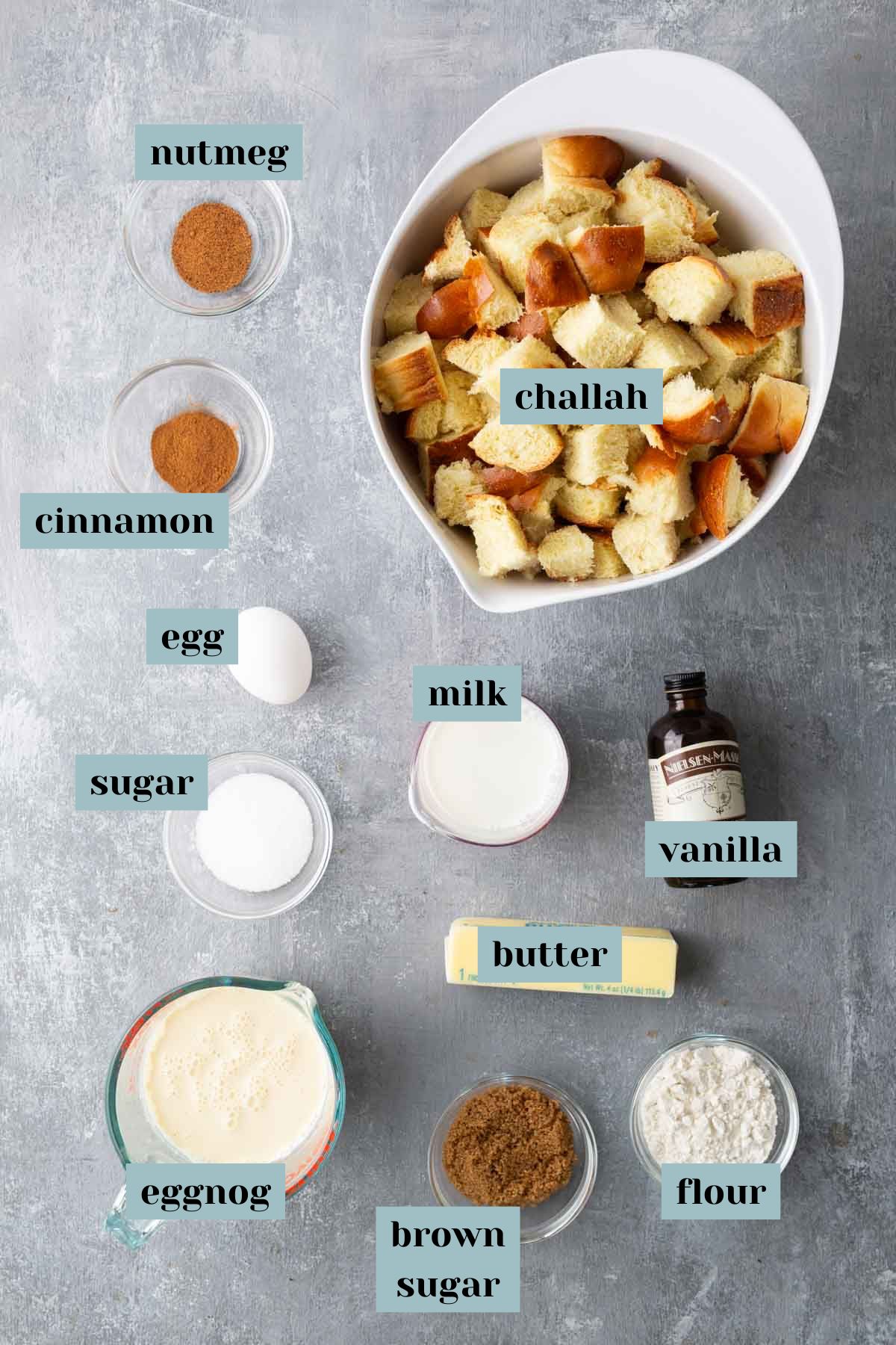 Ingredients laid out for a bread pudding recipe include challah, nutmeg, cinnamon, egg, sugar, milk, vanilla, eggnog, butter, brown sugar, and flour.