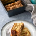 Slices of cinnamon apple bread served on a plate with a fork, with a loaf in the background and a striped cloth.