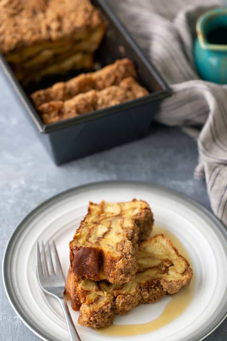 Slices of cinnamon apple bread served on a plate with a fork, with a loaf in the background and a striped cloth.