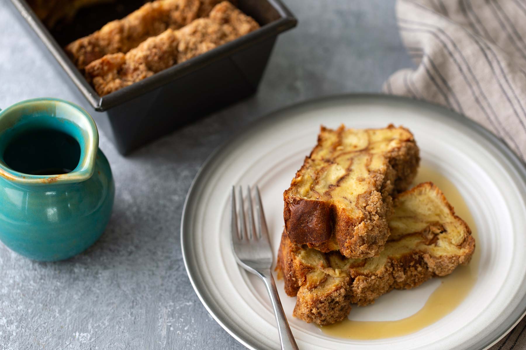 Two slices of cinnamon swirl bread on a plate with syrup, next to a fork and a small green pitcher, with a loaf pan containing more bread in the background.