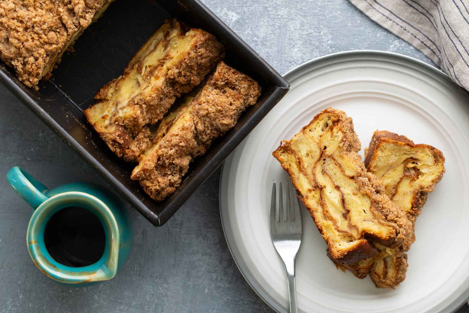 Slices of apple cinnamon bread on a white plate with a fork, next to a loaf pan with more bread and a mug filled with a dark beverage.