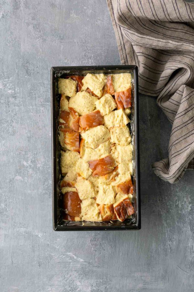 Bread chunks soaked in a yellow batter inside a rectangular baking pan on a gray surface, with a striped cloth on the side.