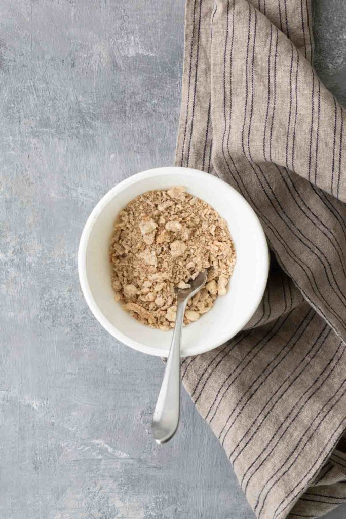 A bowl of dry oatmeal with a spoon, placed on a textured gray surface, next to a striped cloth.