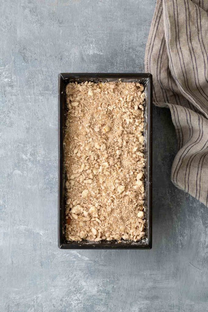 A rectangular baking dish filled with crumbled topping sits on a textured gray surface next to a striped cloth.