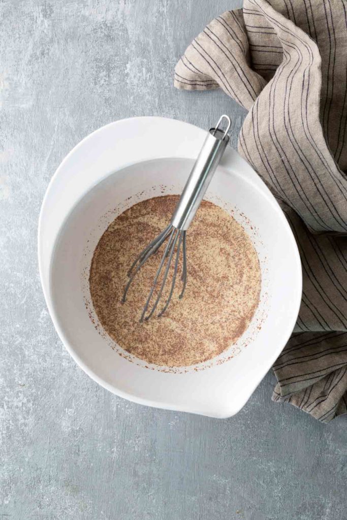 A whisk in a white bowl with a dry mixture, next to a striped cloth on a textured surface.