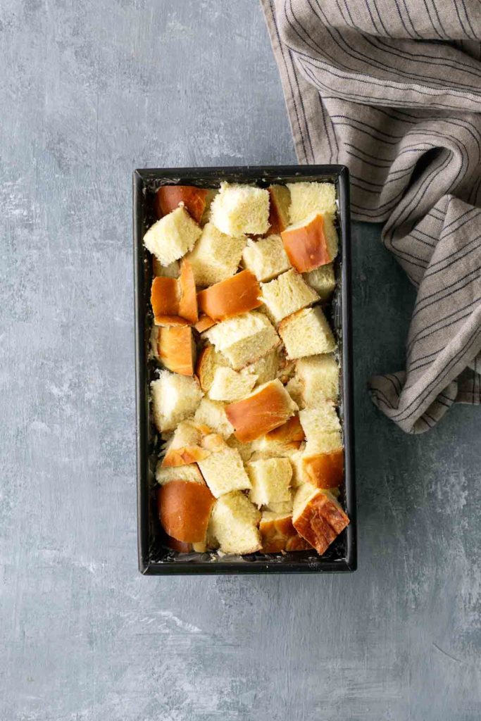 Chopped bread cubes in a rectangular baking pan next to a striped cloth on a gray textured surface.