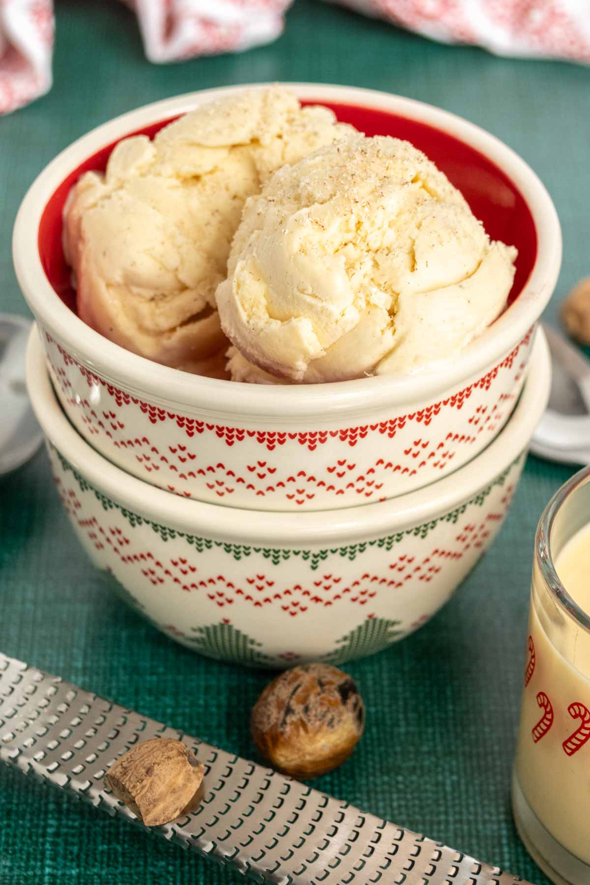 Two scoops of vanilla ice cream in a festive bowl, surrounded by a grater, whole nutmeg, and a glass of creamy beverage on a green surface.