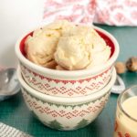 Two scoops of vanilla ice cream in a decorative bowl, with a spoon and a glass of creamy drink nearby on a green tablecloth.