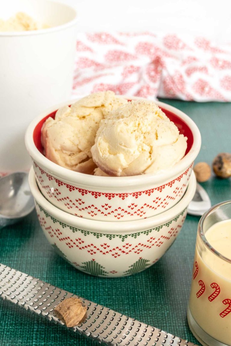 Two scoops of vanilla ice cream in a decorative bowl, with a spoon and a glass of creamy drink nearby on a green tablecloth.