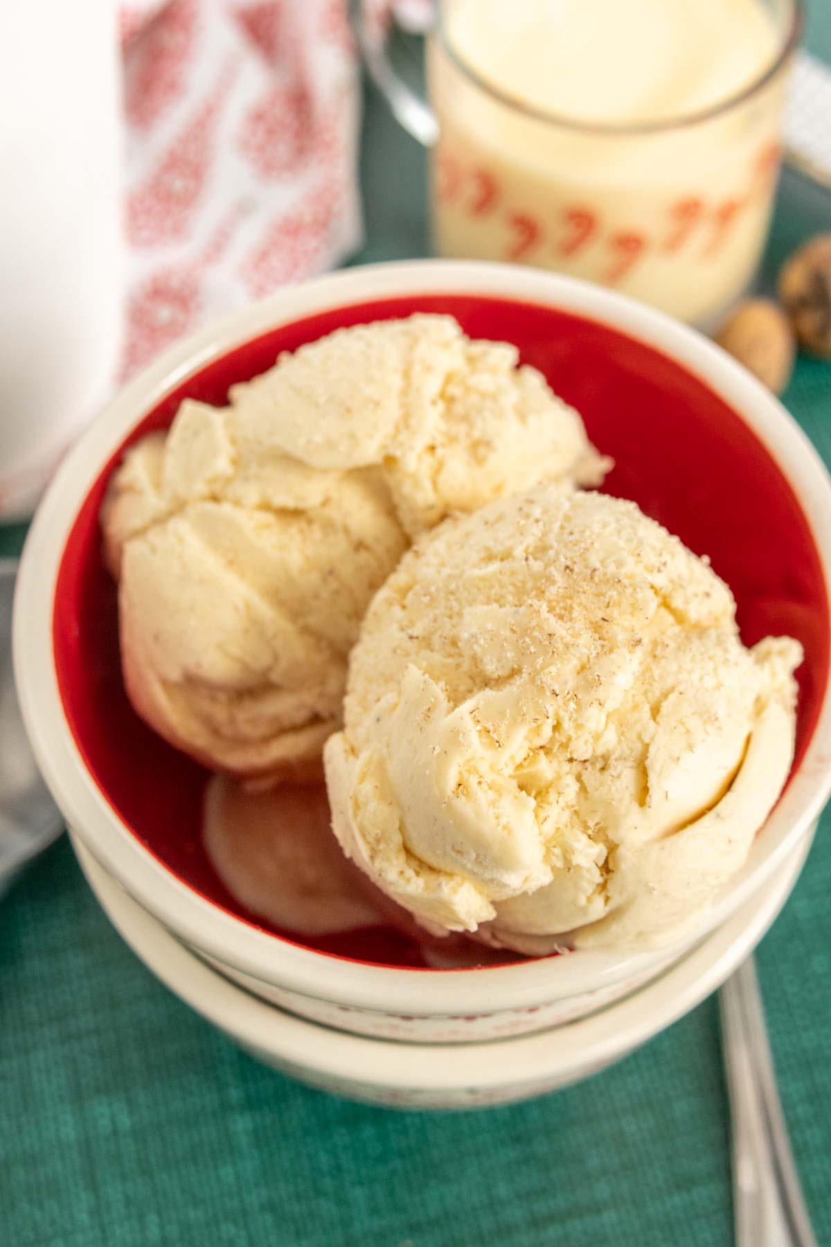 Two scoops of vanilla ice cream in a red bowl, surrounded by a white outer bowl, with a backdrop of a patterned cloth and a measuring cup.