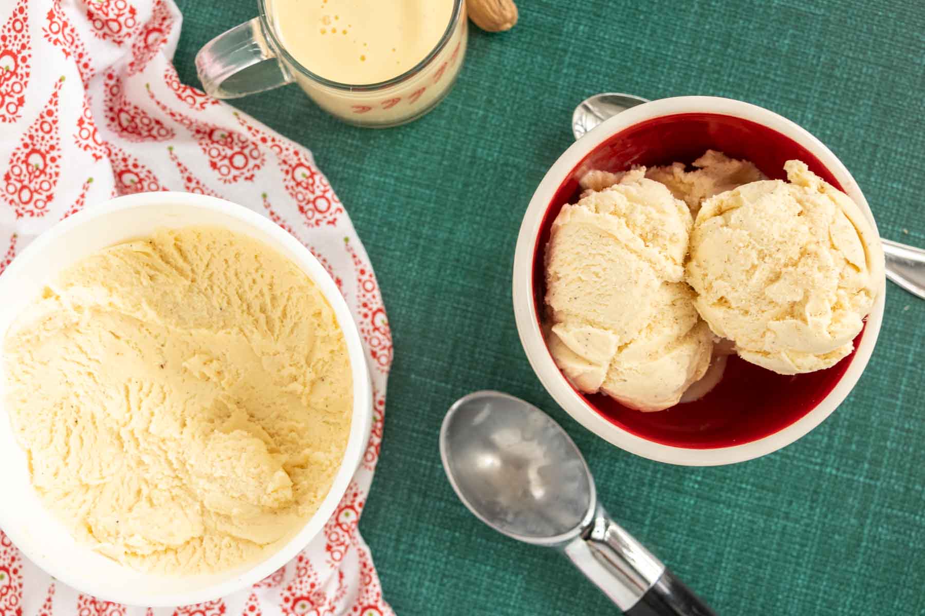 Two bowls of vanilla ice cream with a metal scoop on a green surface. A red-patterned napkin and a glass of light-colored liquid are nearby.