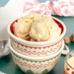 Bowl of eggnog ice cream in a festive patterned dish, with grated nutmeg on top, accompanied by a metal scoop and nutmeg grater.