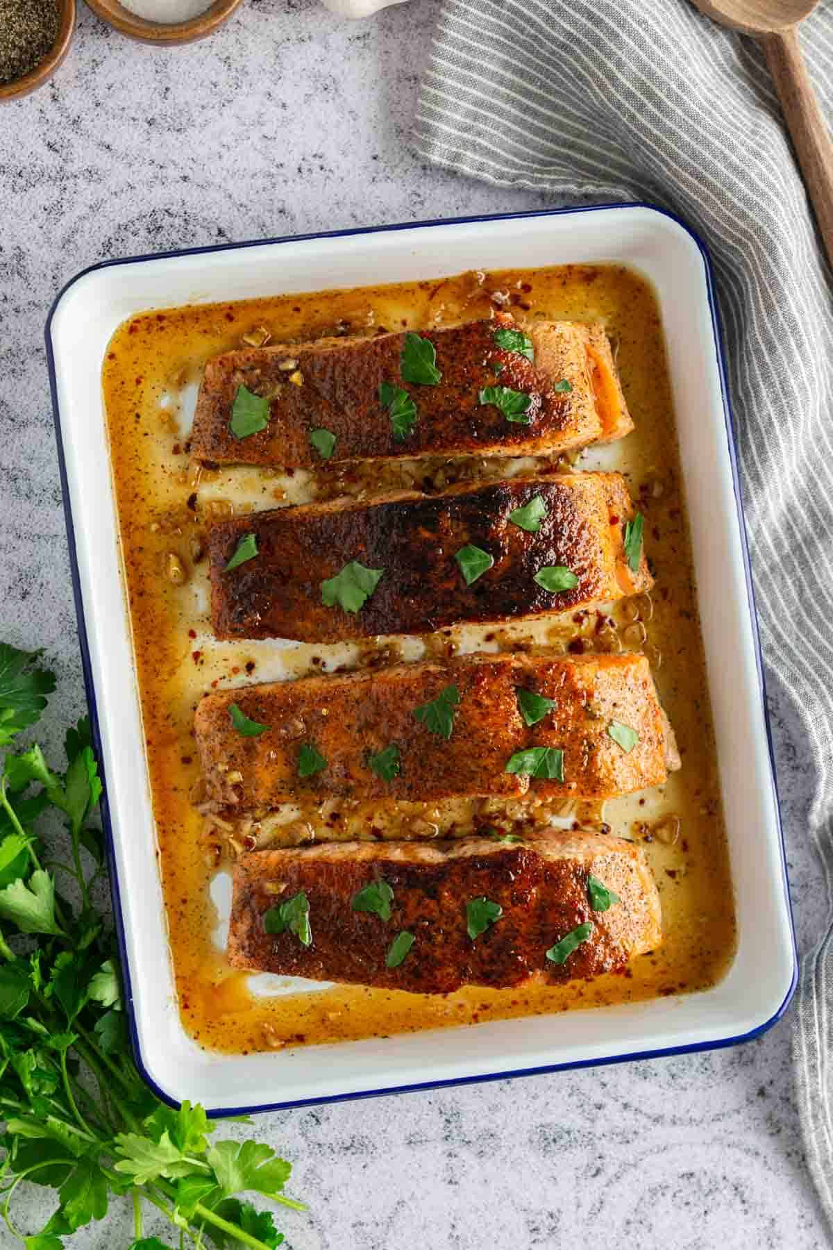 Baked salmon fillets seasoned with herbs and pepper, topped with fresh parsley, in a white baking dish.