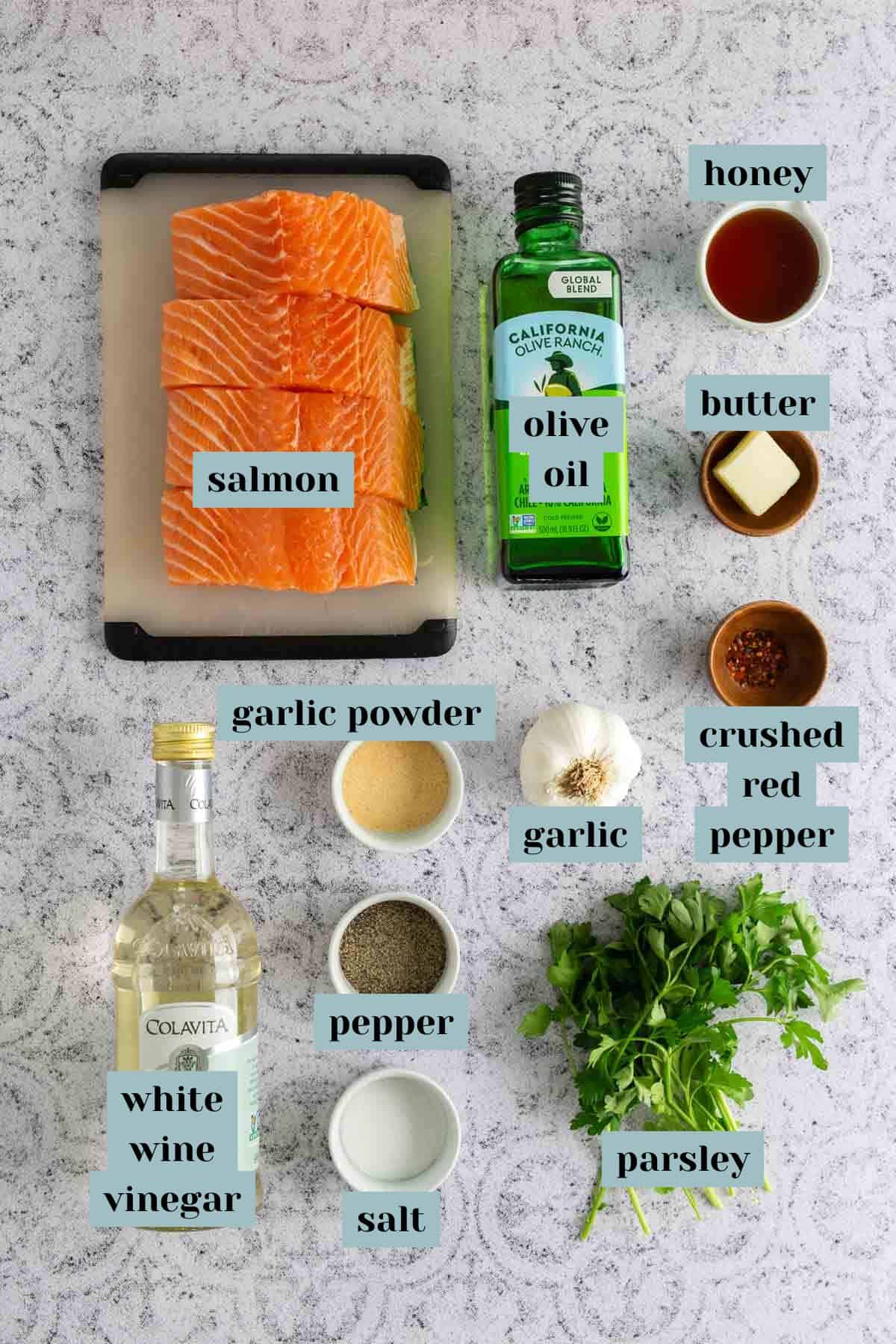 Top-down view of ingredients for a recipe: salmon pieces, olive oil, garlic, honey, butter, crushed red pepper, garlic powder, white wine vinegar, pepper, salt, and parsley on a light surface.