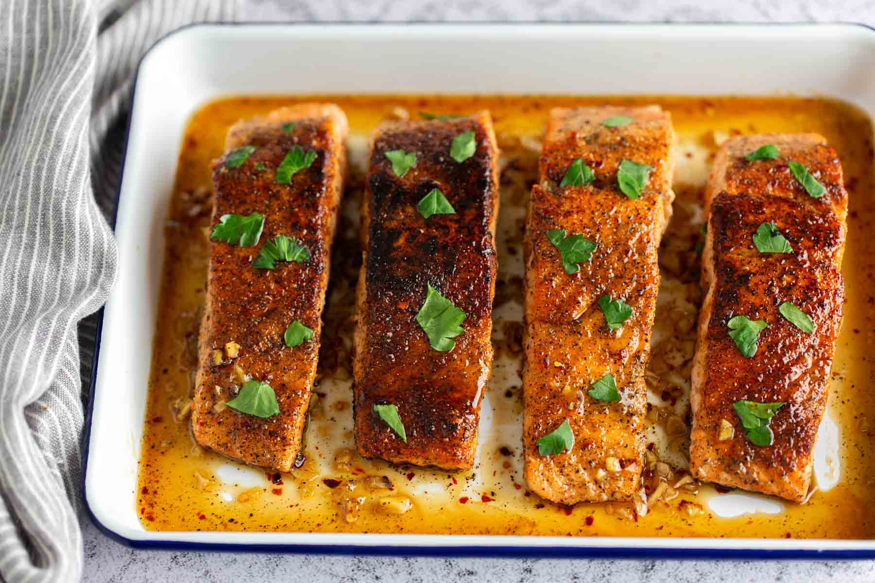 Four seasoned salmon fillets garnished with herbs in a white baking dish.