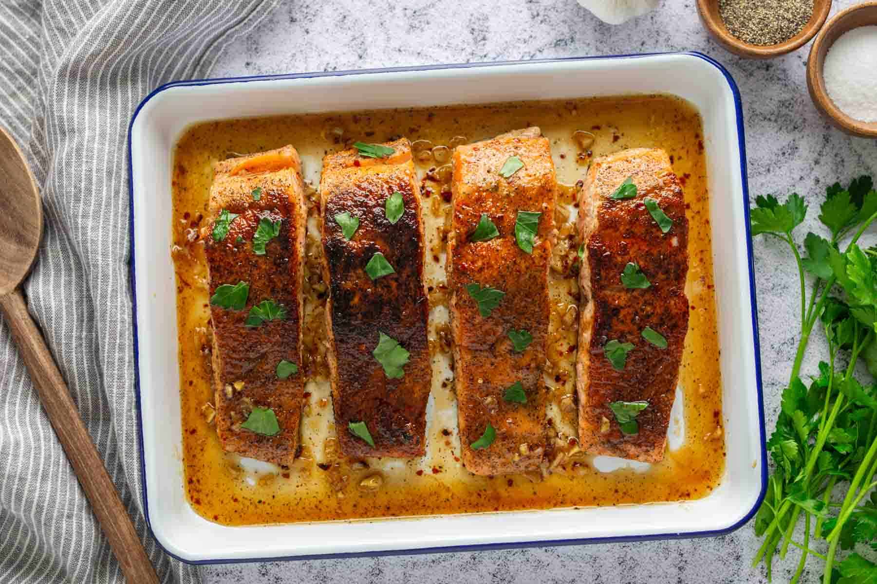 Baked salmon fillets with herbs in a white dish on a textured surface. Parsley and seasoning are nearby.