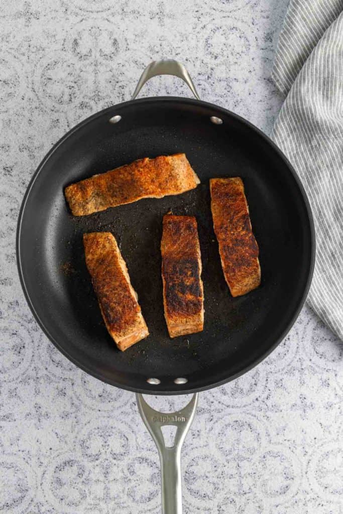 Three pieces of seasoned salmon cooking in a black nonstick frying pan on a gray textured surface with a striped cloth nearby.