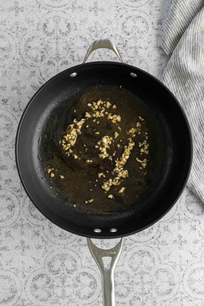 A black frying pan with sautéed garlic in oil, placed on a gray textured surface next to a striped cloth.