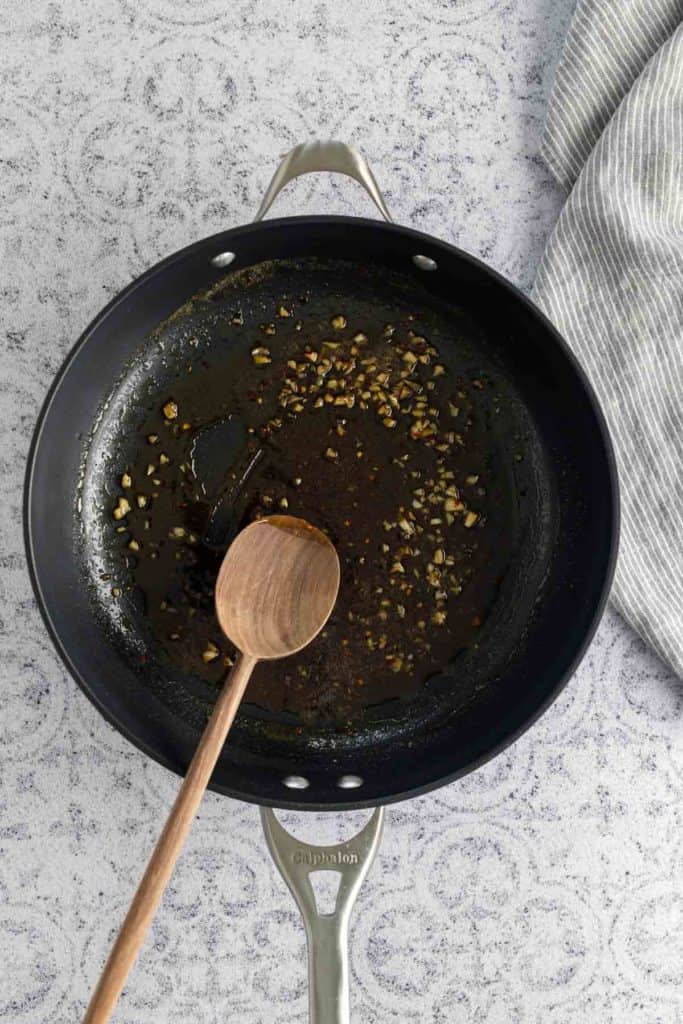 A black frying pan with chopped garlic being sautéed in oil. A wooden spoon rests inside the pan on a patterned countertop, with a striped cloth beside it.