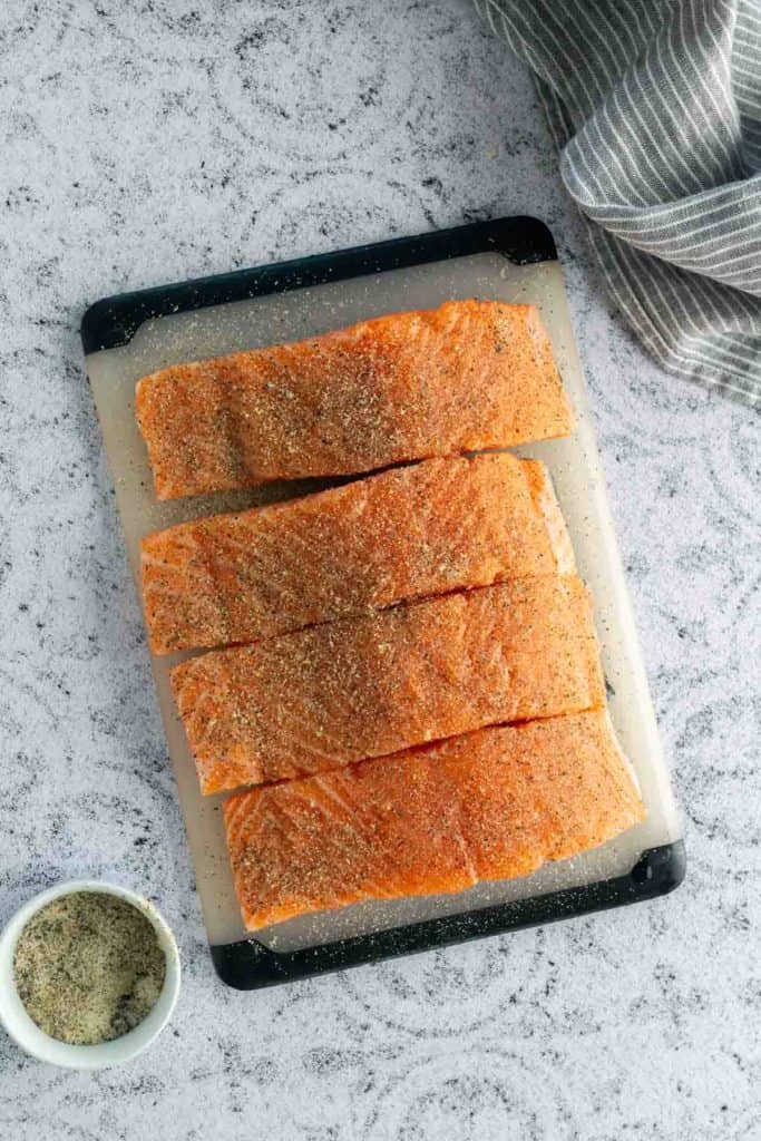 Four seasoned salmon fillets on a cutting board, with a bowl of spices nearby on a speckled countertop.