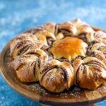 A twisted chocolate bread wreath on a wooden stand, dusted with powdered sugar, set against a blue background.