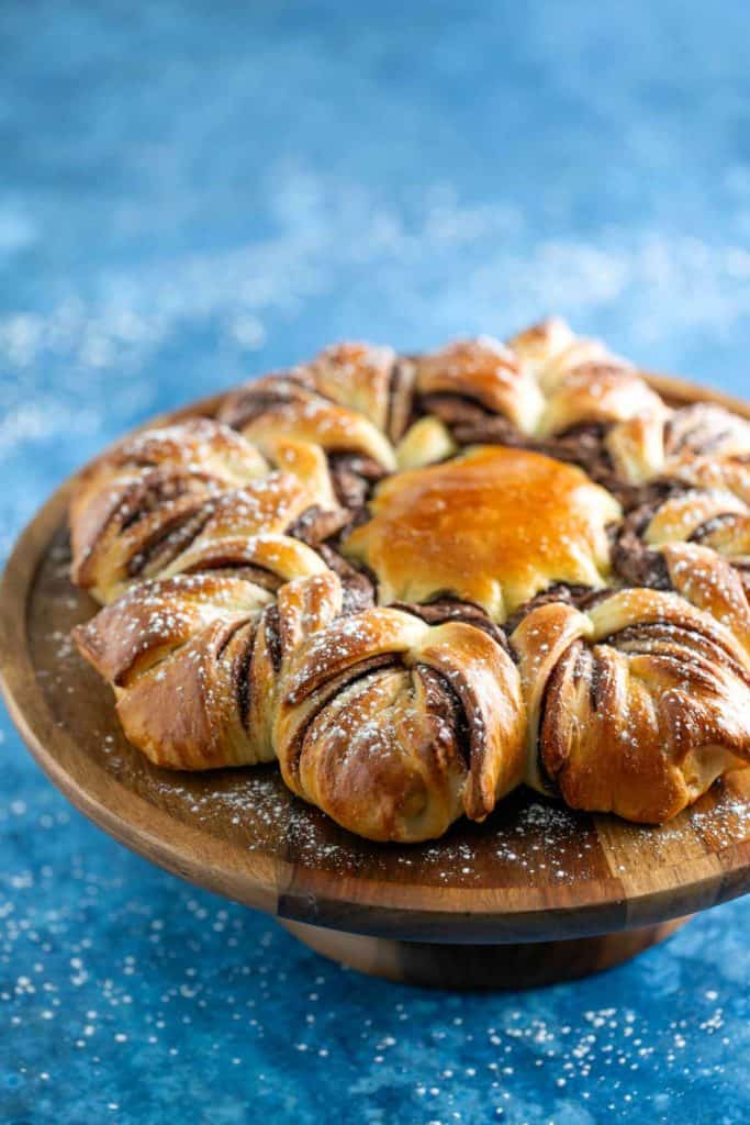 A twisted chocolate bread wreath on a wooden stand, dusted with powdered sugar, set against a blue background.