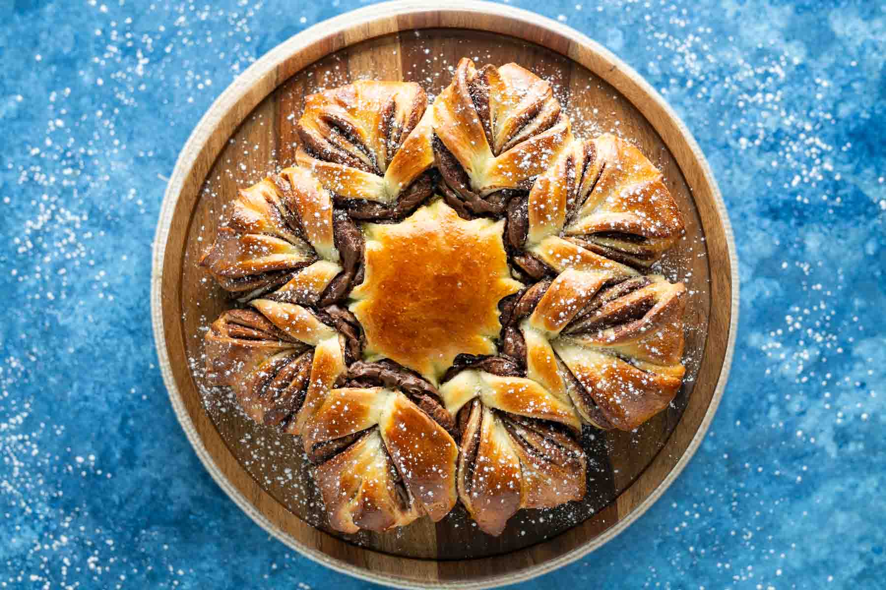 A star-shaped bread with a chocolate swirl design, placed on a wooden serving board. Dustings of powdered sugar are on the bread and blue background.