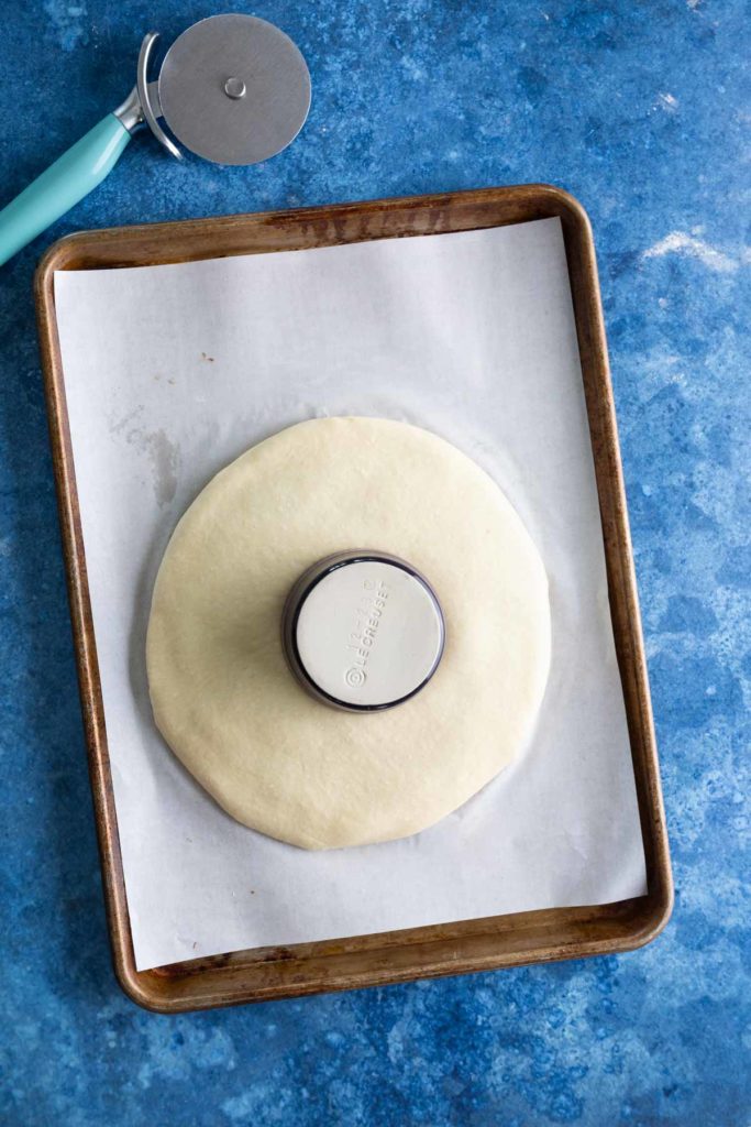 A round piece of dough on a baking tray with parchment paper, with a circular cutter pressing into the center. A pizza cutter is beside the tray on a blue countertop.