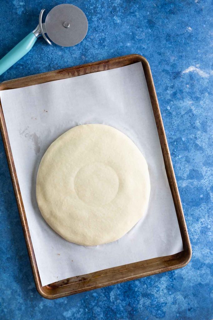 Pizza dough on a parchment-lined baking sheet next to a pizza cutter, on a blue countertop.