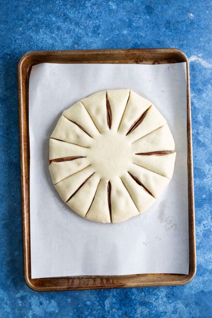 Unbaked circular pastry with radial cuts sits on parchment-lined baking sheet placed on a blue surface.