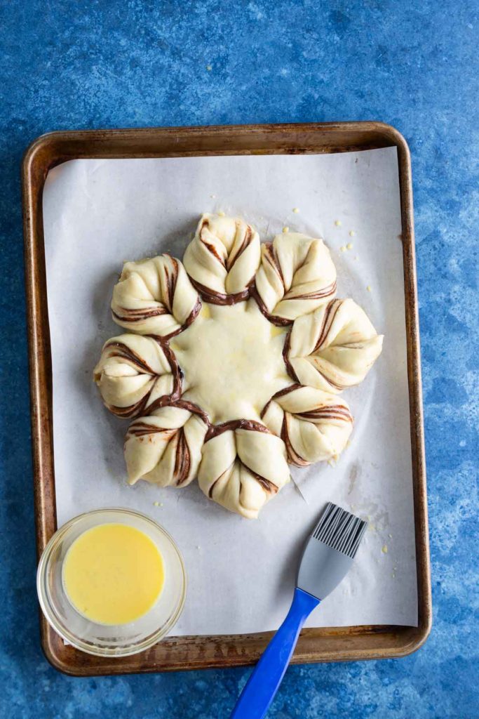 An unbaked star-shaped pastry on a parchment-lined baking sheet with a bowl of egg wash and a blue pastry brush beside it.