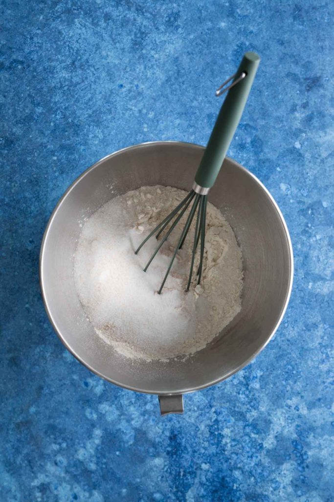 A whisk stands in a mixing bowl containing flour and sugar, set on a blue surface.