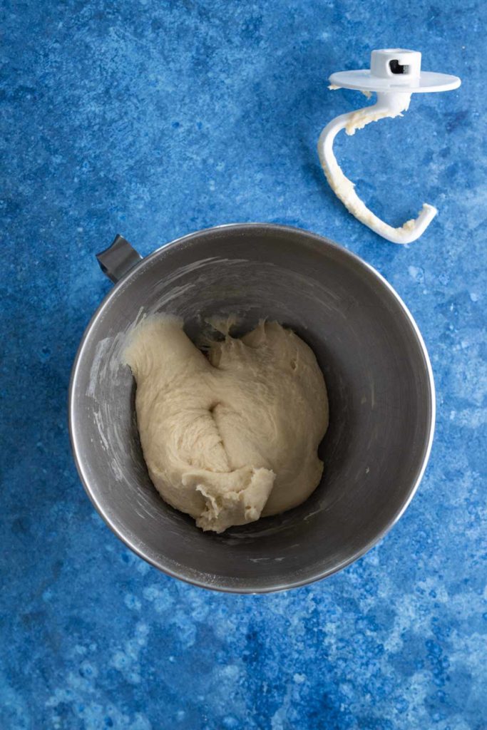 Dough in a metal mixing bowl on a blue surface, with a dough hook attachment nearby.