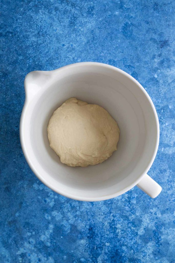 A ball of dough in a white mixing bowl on a blue textured surface.