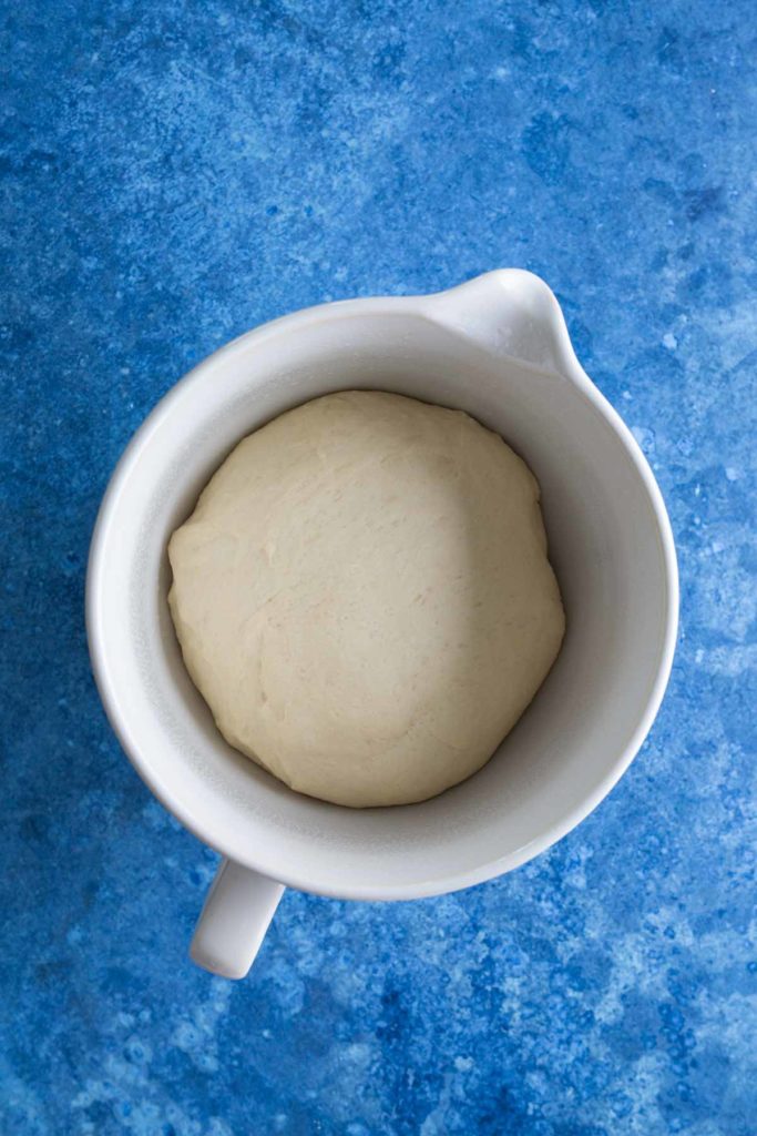 A ball of dough rests in a white bowl against a blue background.