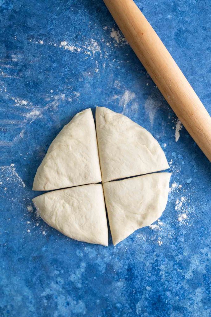 A round piece of dough split into four equal parts on a blue surface, with a wooden rolling pin nearby.