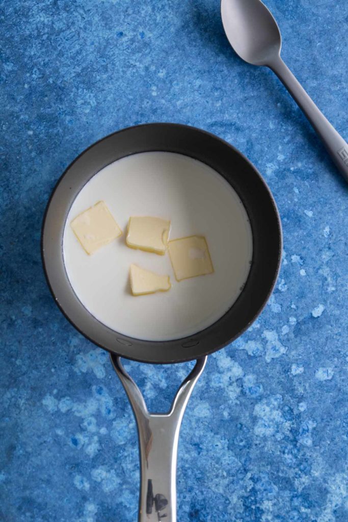 A saucepan with milk and butter pieces on a blue countertop, next to a spoon.