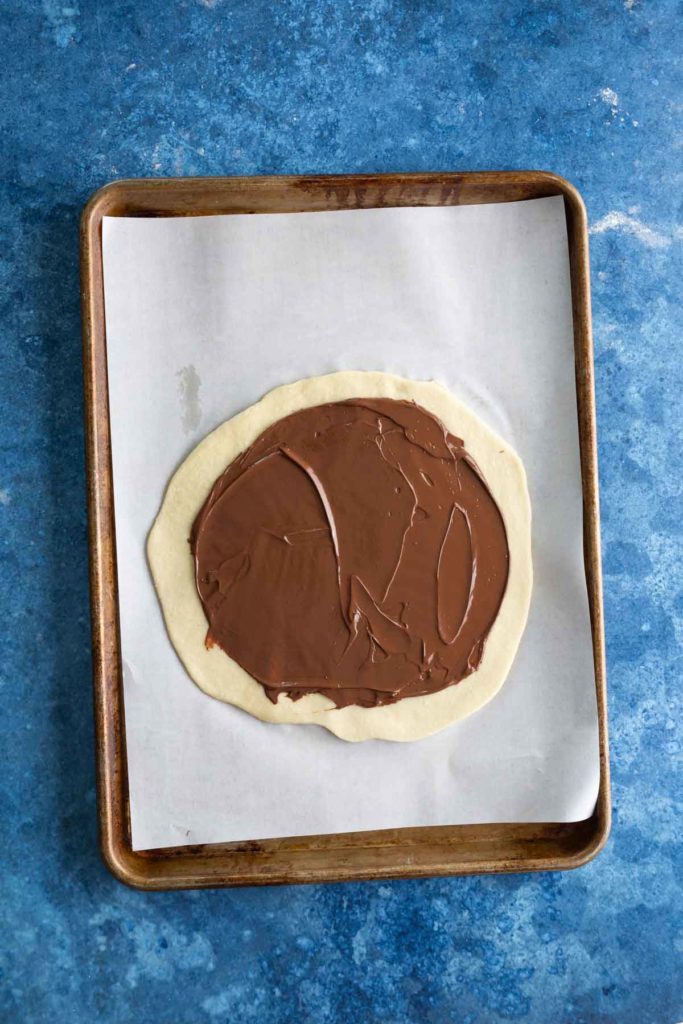 An uncooked dough circle topped with chocolate spread on a parchment-lined baking sheet.