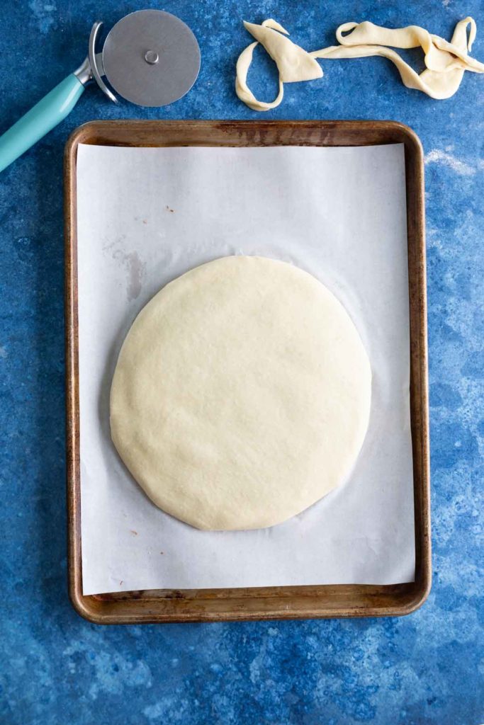 A round pizza dough on a parchment-lined baking sheet next to a pizza cutter and dough scraps on a blue countertop.