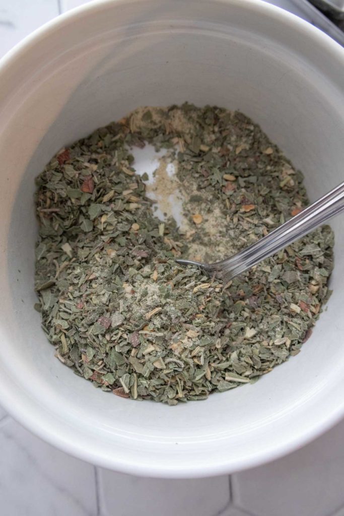 A white bowl containing a mixture of dried herbs with a metal spoon inside.