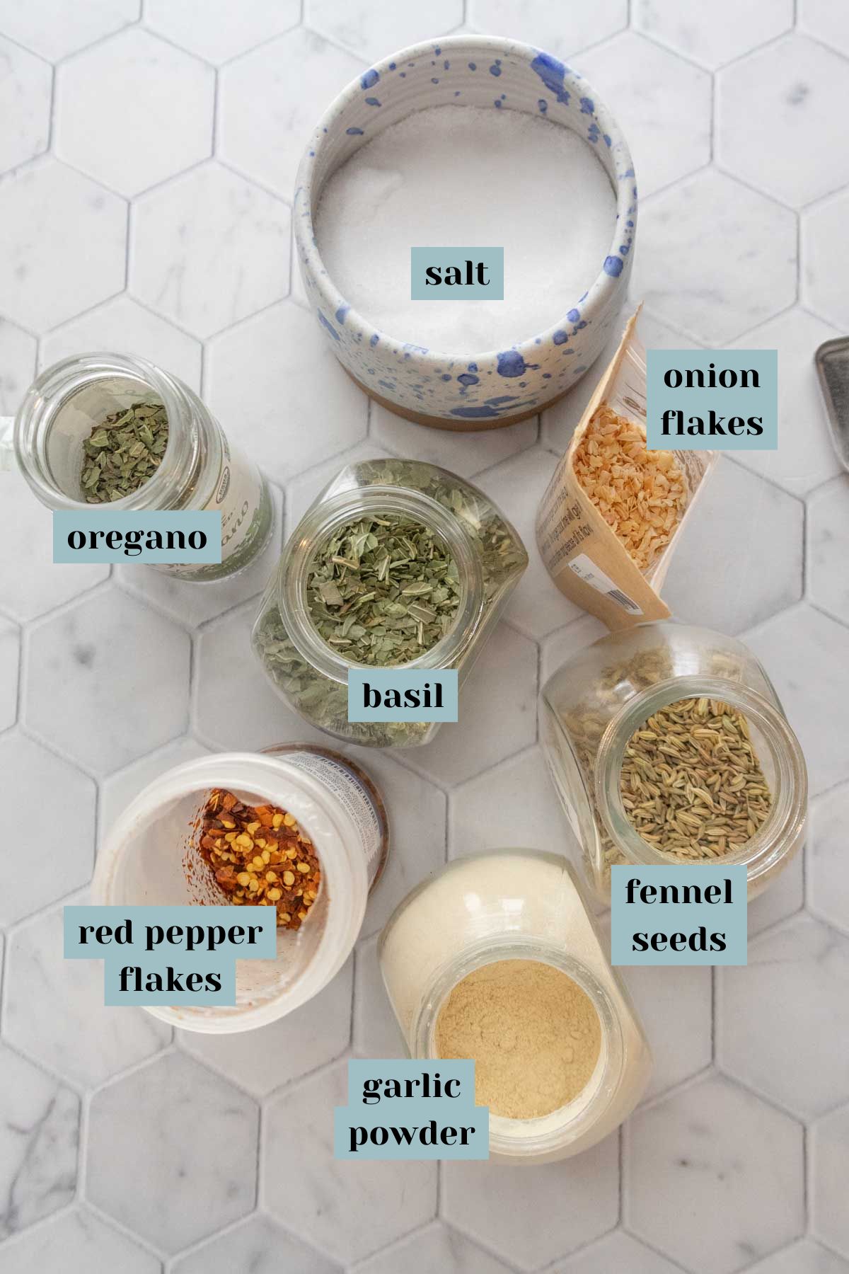 Six spice jars on a hexagonal tile surface, labeled: oregano, basil, salt, onion flakes, red pepper flakes, fennel seeds, garlic powder.