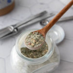 A wooden spoon holds a portion of greenish seasoning mix above an open glass jar on a hexagon pattern countertop.