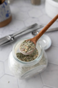 A wooden spoon holds a portion of greenish seasoning mix above an open glass jar on a hexagon pattern countertop.