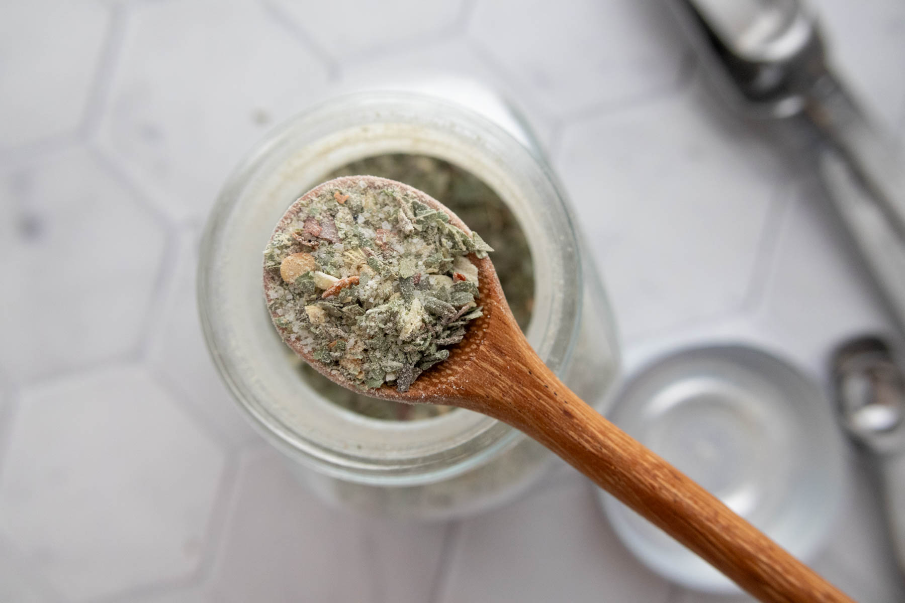 A wooden spoon holds a portion of a coarse, greenish herbal mixture over an open jar on a white surface.